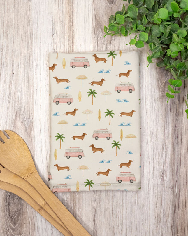 Patterned towel with dachshunds, vans, and palm trees on a wooden surface with wooden utensils and a plant.