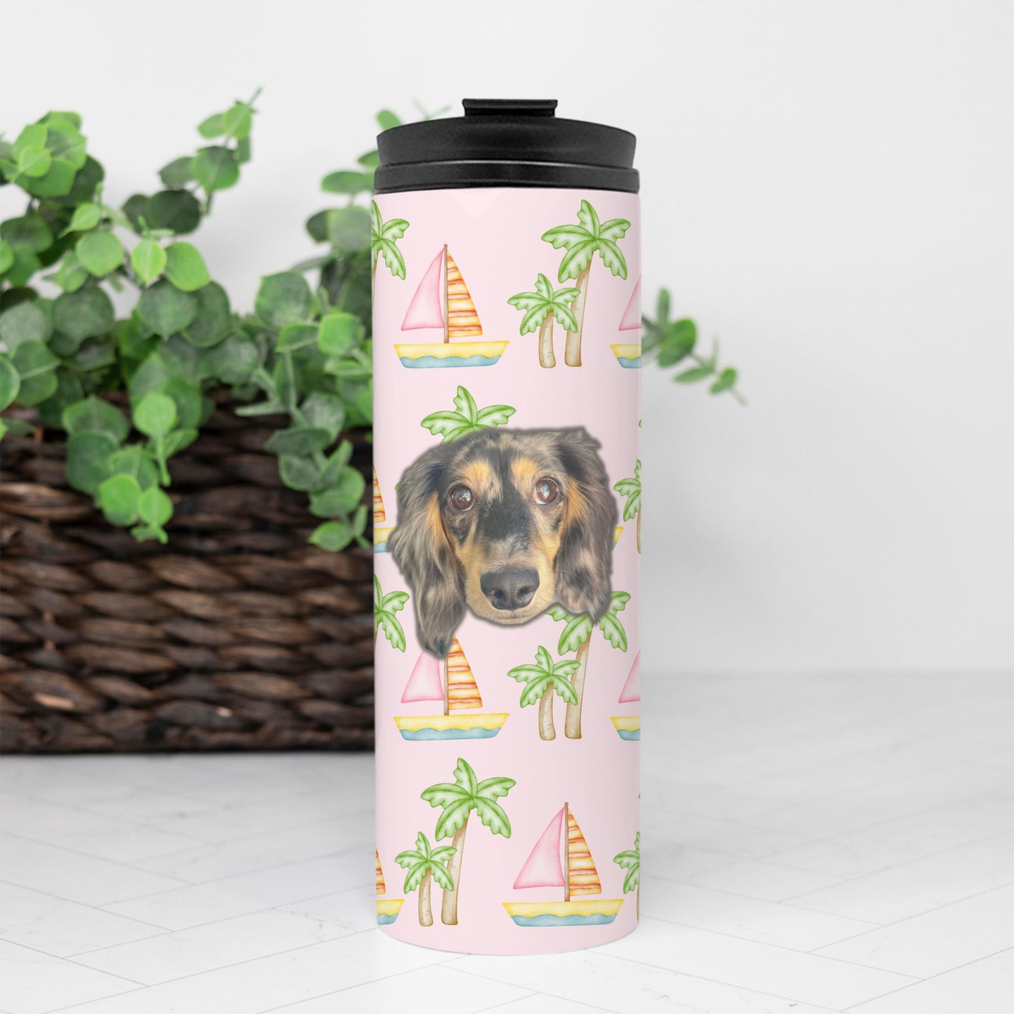 Seafoam thermal tumbler with pink sailboats and green palm trees, personalized with a simple portrait of your pet's face, 16 oz capacity by My Doxie Depot sitting in front of a brown basket and green plant