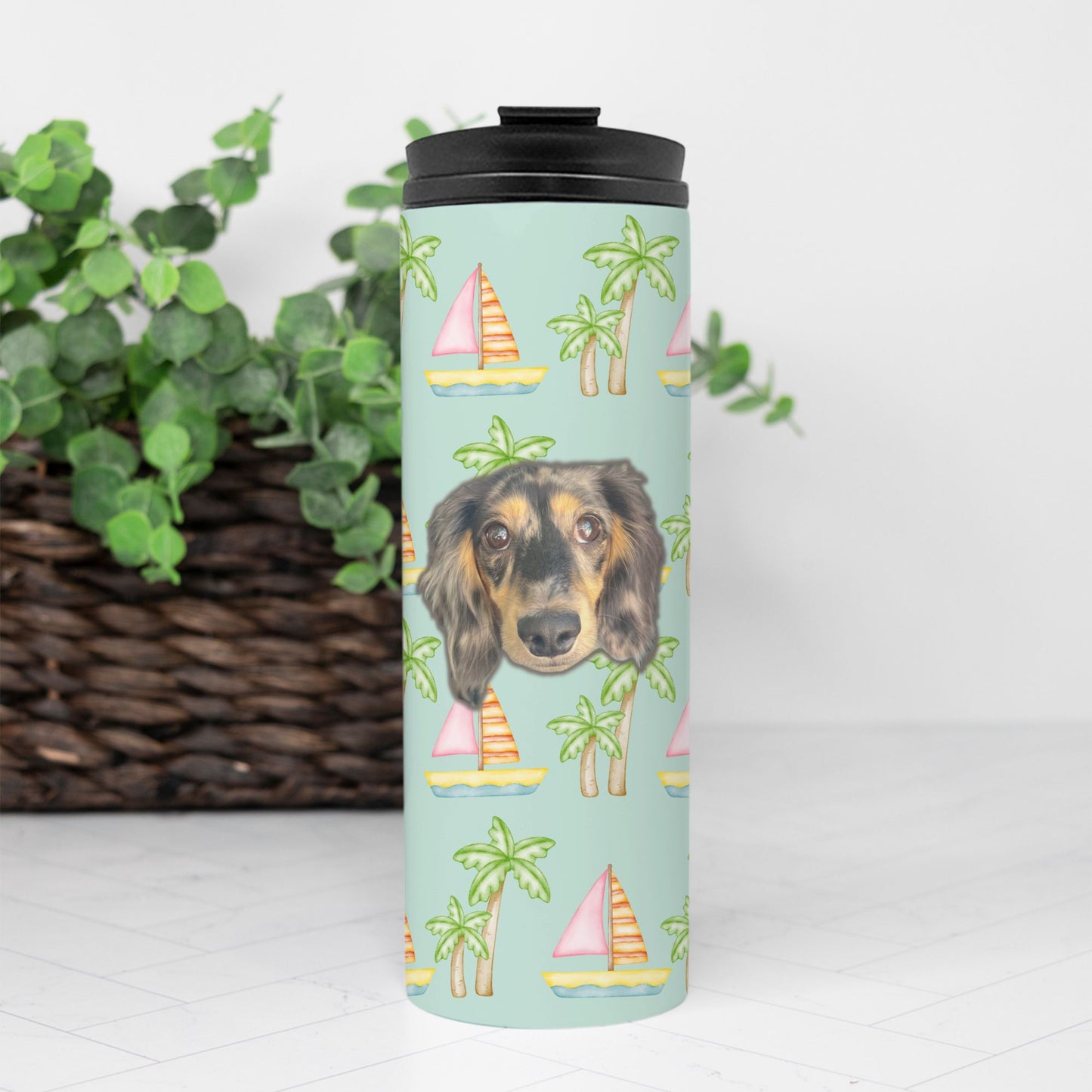 Seafoam thermal tumbler with pink sailboats and green palm trees, personalized with a simple portrait of your pet's face, 16 oz capacity by My Doxie Depot in front of a brown basket and a green plant